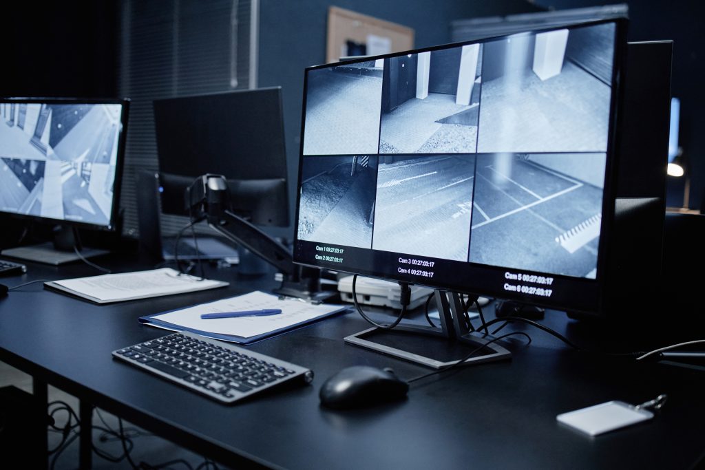 Empty security control room showing modern surveillance workstation with multiple monitors displaying live CCTV footage of building corridors and parking areas, keyboard and documents on desk